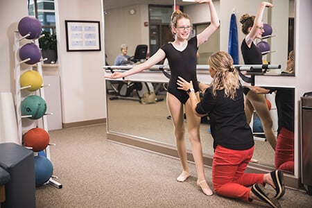 Therapist kneeling on the floor to correct the posture of young ballerina.