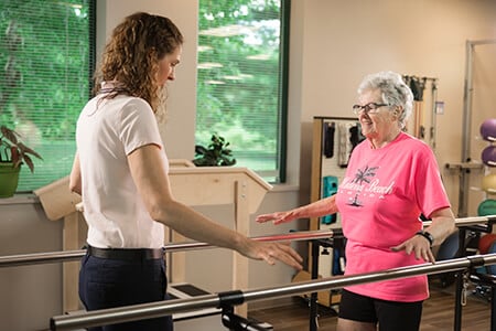 Older female patient in bright pink shirt using parallel bars to practice walking.