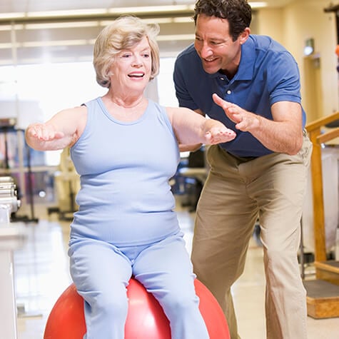 Therapist spotting an older female sitting on a large balance ball with her arms stretched in front of her.