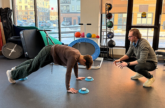 Atlee is holding a plank on the floor as Tom is squatting down beside him
